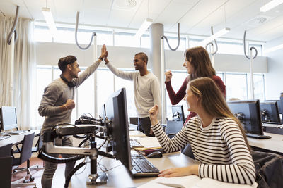 Happy male and female students celebrating success while using computers at library in university