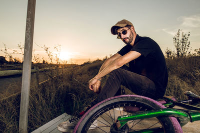 Portrait of man sitting with bicycle on field against sky during sunset