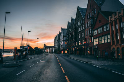 Road by buildings against sky during sunset