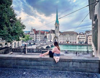 Woman sitting by buildings against sky in city