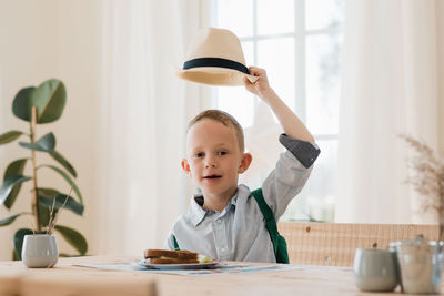 Boy holding his hat up whilst eating his lunch at home smiling