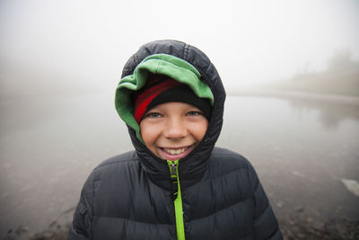 Portrait of smiling boy wearing warm clothing against lake during foggy weather