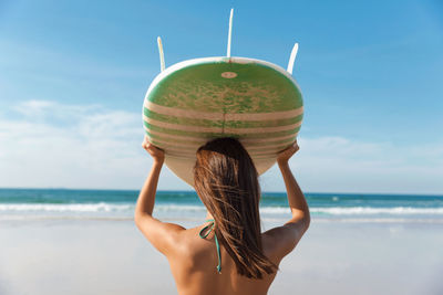 Rear view of young woman holding surfboard while standing at beach against sky