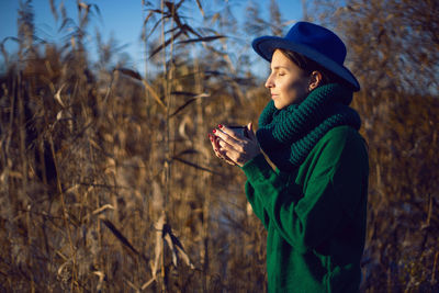 Woman in knitted sweater and blue hat stands bushes sunset and drinks tea from an iron thermos mug