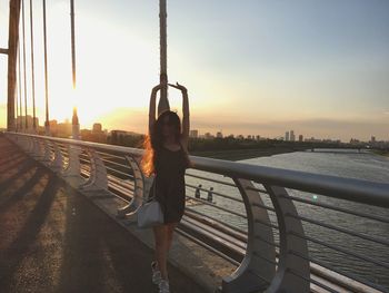 Full length of woman with arms raised at bridge over river during sunset