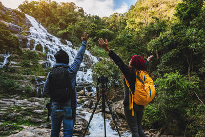 Rear view of friends with arms raised standing by waterfall and camera in forest