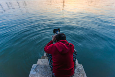 Rear view of man photographing through camera while sitting on steps at lake