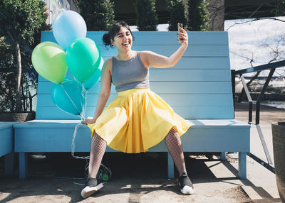 Happy young woman taking selfie with balloons while sitting on bench