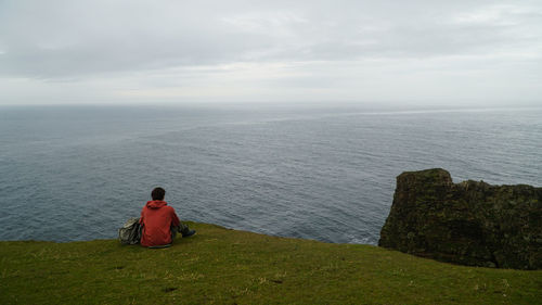 Rear view of man sitting cliff by sea against sky