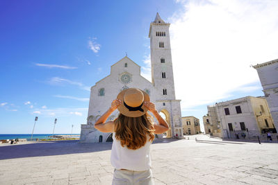 Back view of beautiful tourist woman holding her hat in trani with its cathedral in apulia, italy.