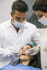 Male dentist in mask and gloves examining teeth of pretty patient with help of female assistant at clinic