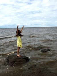 Side view of girl with arms raised standing on rock formation in sea