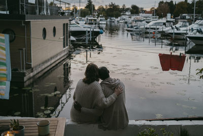 Rear view of couple with arm around sitting on wall while looking at marina