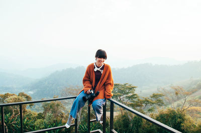 Young asian woman wearing casual clothes holding camera sit on fence at mountain top