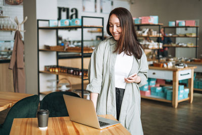 Adult business woman with long hair in stylish shirt working on laptop using mobile phone in cafe