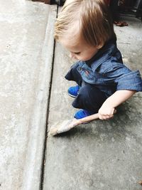 High angle view of boy holding paintbrush while crouching on footpath