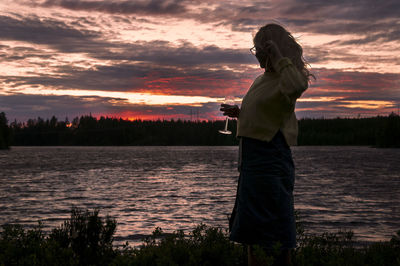 Woman standing by lake against sky during sunset