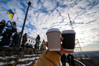 Cropped hand of man holding coffee cups against sky