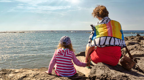 Mother and little daughter sitting on rocky seacoast holding hands. rear view. 