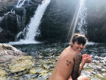 Portrait of smiling shirtless woman sitting by waterfall
