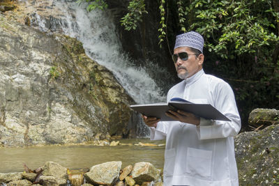 Man reading book while standing against waterfall