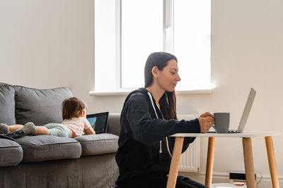 Mother using laptop while daughter watching digital tablet on sofa at home