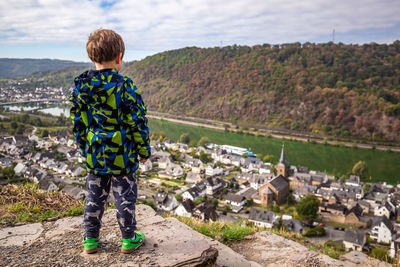 Rear view of boy standing on cliff by house and river in town