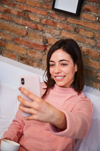 Young woman taking a selfie with mobile phone while lying on bed at home