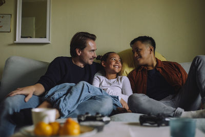 Smiling gay men spending leisure time with daughter while sitting on sofa at home