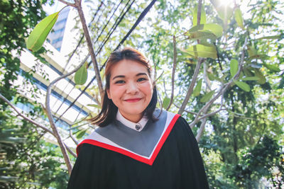 Portrait of smiling young woman wearing graduation gown against tree