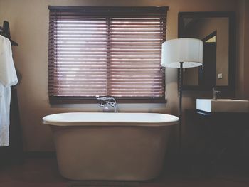 Bath tub by lamp against window in bathroom