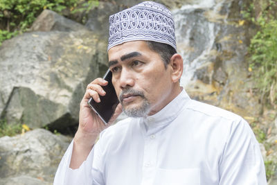 Thoughtful man talking on phone while sitting against waterfall