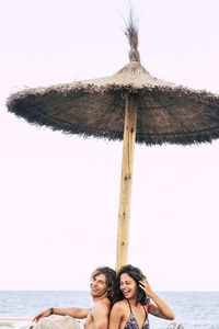 Happy couple sitting under parasol at beach against clear sky