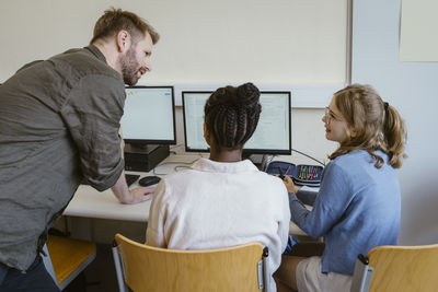 Male teacher discussing with female students sitting on chairs in computer class at school