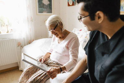 Elderly woman looking in mirror while sitting by male caregiver at bedroom