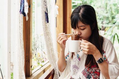 Young woman drinking coffee while sitting by window at home