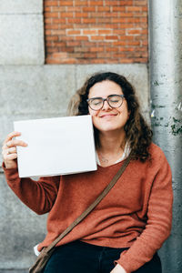 Smiling young woman with eyes closed holding blank paper while sitting against wall