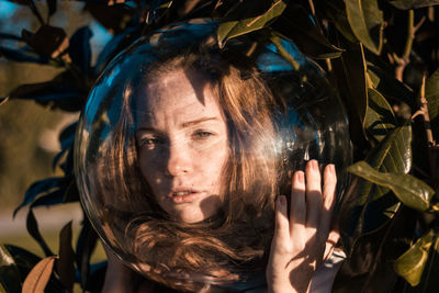 Close-up portrait of young woman wearing glass ball on head against leaves