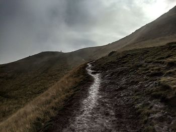 Pathway on mountain against cloudy sky during sunny day