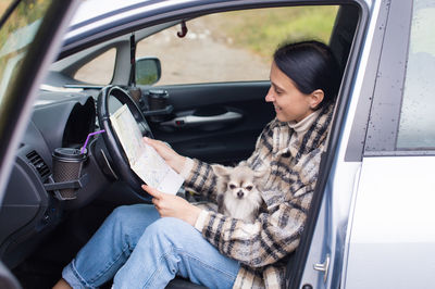 A cute girl is sitting in a car behind the wheel, holding a chihuahua and a road map, smiling