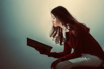 Side view of woman reading book while sitting against wall