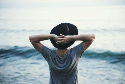 Rear view of young woman wearing hat with hands behind head against sea
