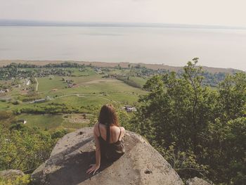 Rear view of woman sitting on rock by sea against sky