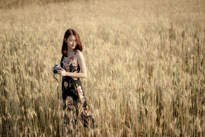 Young woman holding camera while standing amidst crops on agricultural field