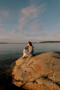 Woman sitting on rock by sea against sky during sunset