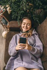 Young beautiful woman lies near christmas tree in living room on sofa, smiling and using phone,
