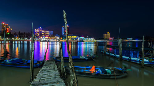 Boats moored on river by illuminated city against sky at night