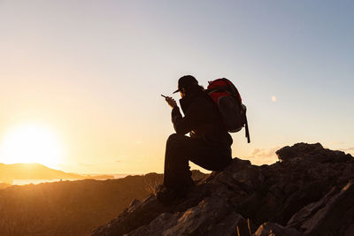 Side view of unrecognizable male hiker sitting on rock in highlands and speaking on cellphone while admiring sunset