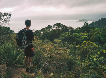 Rear view of man with backpack looking at sea while standing on mountain against cloudy sky