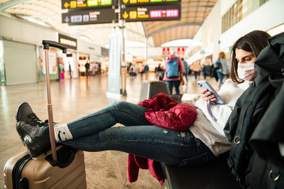 Full body female in casual clothes and face mask holding legs on suitcase and browsing cellphone while waiting for flight in airport terminal during pandemic